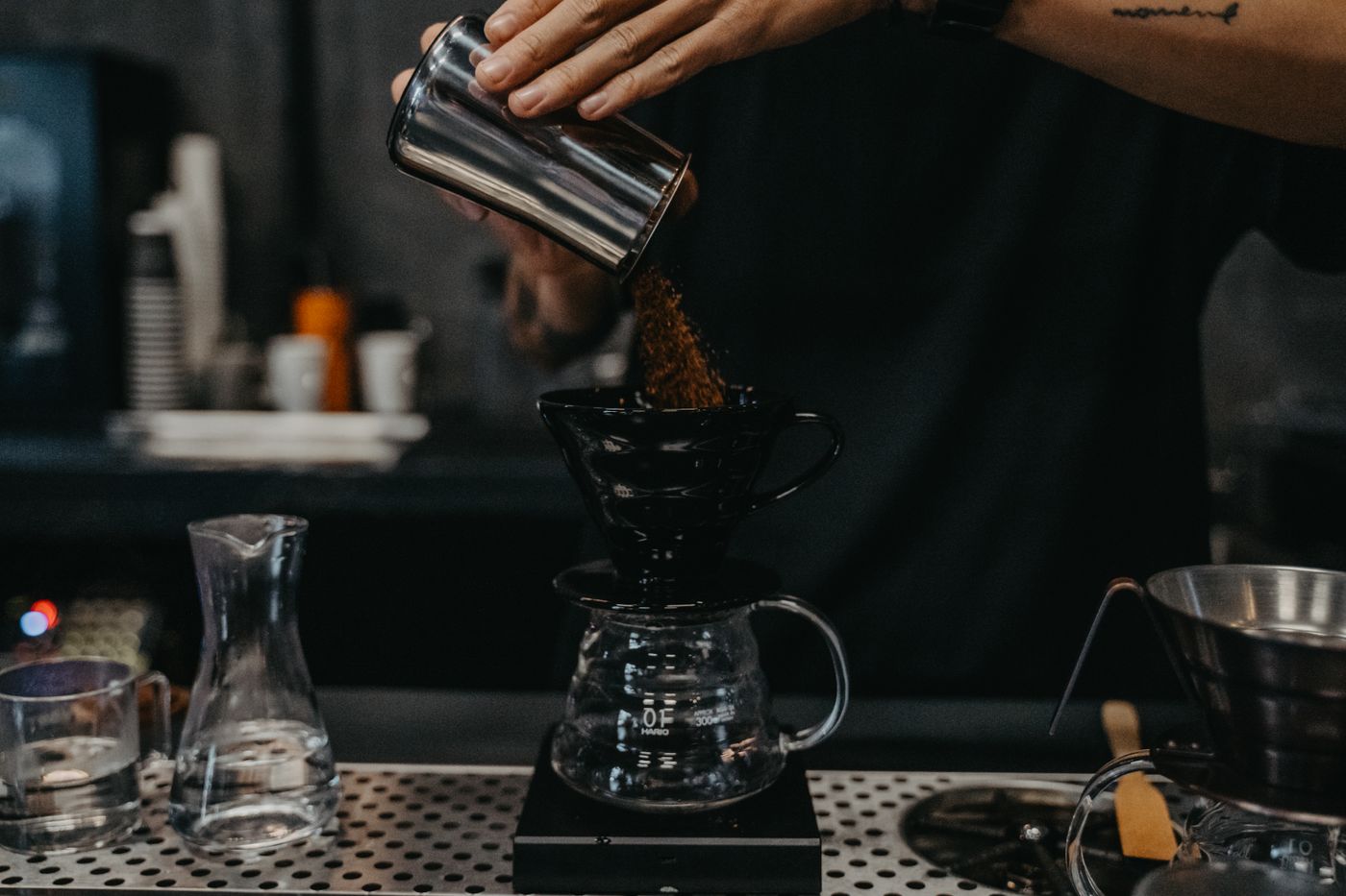 A barista dosing freshly ground coffee into a Hario V60 dripper over a glass server on a brew scale